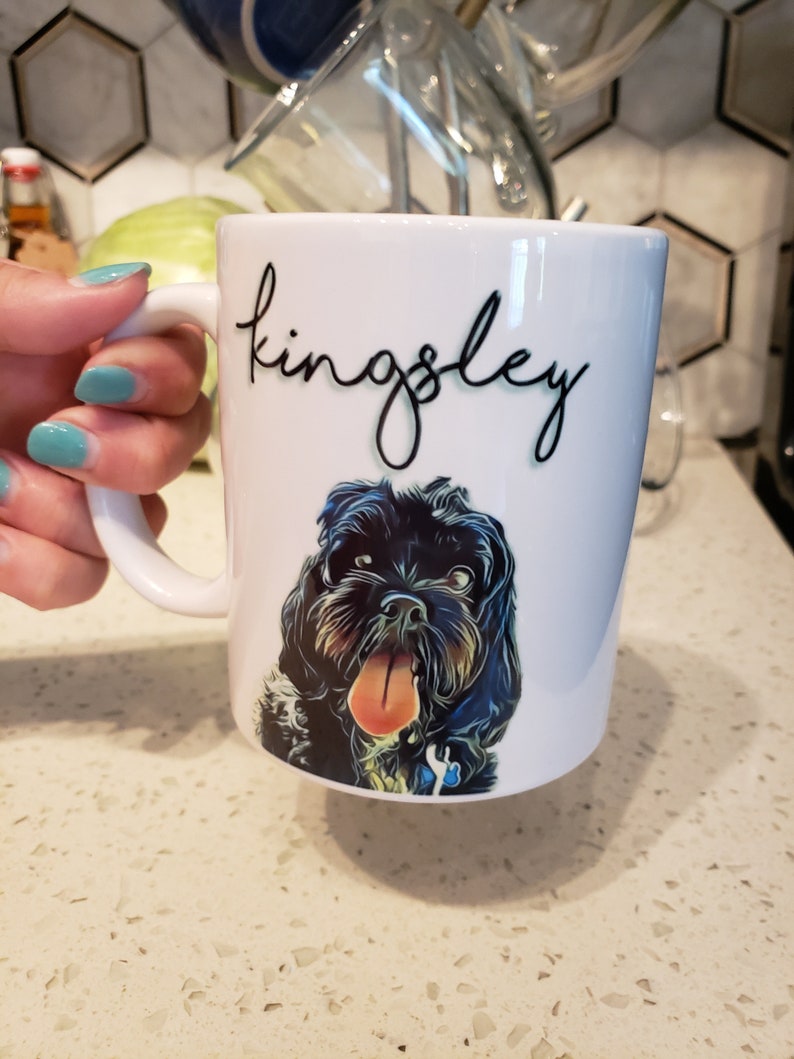 May include: White ceramic mug with a black and white illustration of a dog with its tongue sticking out. The mug has the name "Kingsley" written in black cursive on the side.