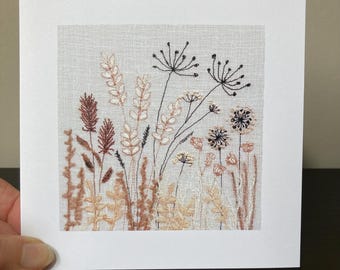 May include: A handmade embroidered card featuring a detailed floral design. The artwork showcases various wildflowers and grasses in shades of brown, beige, and black, set against a light gray background. The card is presented on a white square.