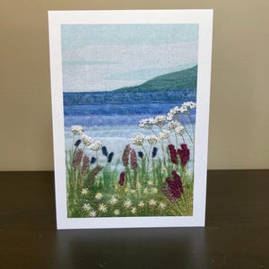 May include: A handmade card featuring an embroidered landscape with a blue sea, green hills, and a field of white and purple flowers.