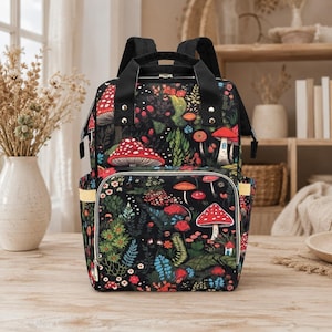Puede incluir: Mochila negra con un colorido estampado de setas y flores. La bolsa tiene un asa superior, correas ajustables y múltiples bolsillos. El diseño presenta setas rojas, blancas y naranjas, follaje verde y pequeñas flores.