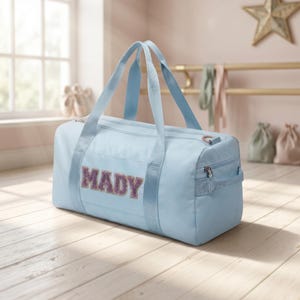 Puede incluir: Una bolsa de deporte azul claro con el nombre "MADY" en letras moradas brillantes. La bolsa tiene dos asas y un cierre de cremallera. La bolsa está sobre un suelo de madera.