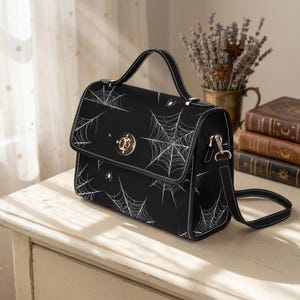 Puede incluir: Bolso de mano negro con diseño de telaraña blanca. Tiene un asa superior y una correa de hombro desmontable. El cierre es dorado y las costuras son blancas. El bolso está sobre una superficie blanca.