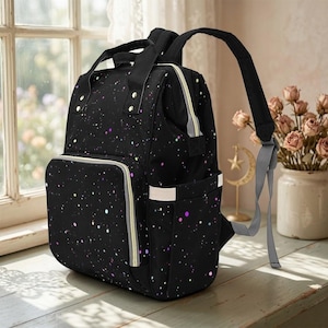 May include: Black diaper bag with a colourful speckled pattern. The bag has a front pocket, side pockets, and a top handle. It features a zip closure and adjustable shoulder straps. The bag is on a wooden surface near a window.