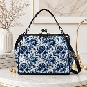 May include: A structured handbag featuring a white and blue floral pattern with a kiss-lock closure. The bag has a black frame, a black top handle, and a detachable black shoulder strap. The exterior displays a repeating dark blue peony design.