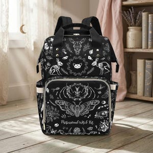 A black diaper bag backpack featuring a white witchy motif print sits upright on a light wooden floor.