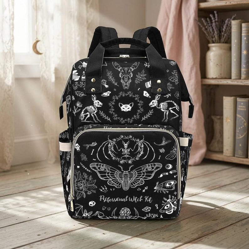 A black diaper bag backpack featuring a white witchy motif print sits upright on a light wooden floor.