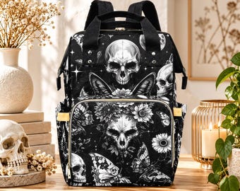 Puede incluir: Una mochila negra con un estampado gráfico blanco de calaveras, mariposas y flores. La bolsa incluye un asa superior, correas ajustables para los hombros, un bolsillo frontal con cremallera y detalles en tono dorado en los laterales y cremalleras.
