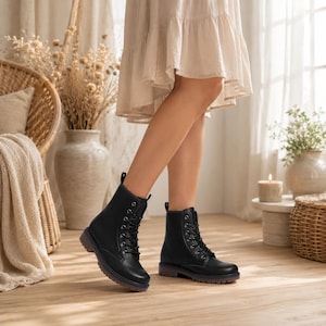 May include: Black lace-up ankle boots with a rounded toe and a low, dark sole. The boots have a matte finish and are worn with a cream-colored dress. The boots are a classic style, suitable for various outfits.