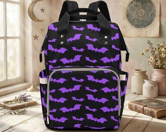May include: A black backpack featuring a repeating pattern of purple bats. The bag includes a large main compartment, a front zippered pocket, two side pockets, and black shoulder straps with silver-toned metal hardware accents.