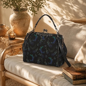 Puede incluir: Un bolso de mano negro con un estampado de lunas crecientes y ramas de hojas en tonos azules y verdes. El bolso cuenta con una estructura rígida, un cierre metálico de boquilla con detalles de filigrana, un asa superior y una correa extraíble.