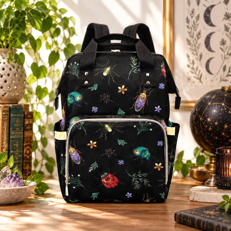 May include: A black backpack featuring a colourful pattern of various insects, including a red ladybird, beetles, and small green plants. The bag has a large main compartment, a front zipped pocket, two top handles, and adjustable shoulder straps.
