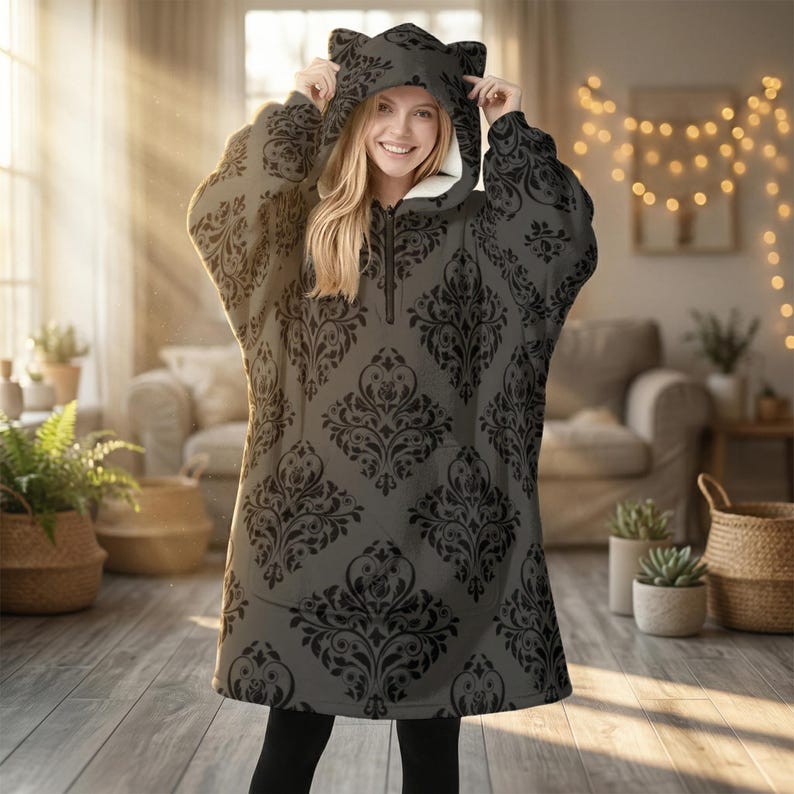 May include: A gray hooded blanket with a black damask pattern. The blanket has a zipper closure and cat ears on the hood. The interior of the hood is white. The person is wearing black leggings.