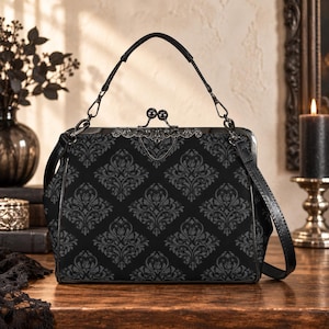 May include: A black handbag featuring a dark grey damask pattern, a metal kiss-lock closure with ornate detailing, a top handle, and a detachable shoulder strap. The bag has a structured frame and a rectangular shape with rounded bottom corners.