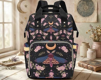 May include: A black backpack featuring a repeating pattern of moths, crescent moons, and pink flowers. The bag has black top handles, adjustable shoulder straps, a front zippered pocket, and side pockets with light-colored trim accents.