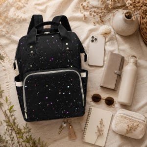 May include: A black backpack with a galaxy design, speckled with colourful dots. The image also shows a phone, wallet, sunglasses, notebook, water bottle, and a small purse, all arranged on a neutral surface.
