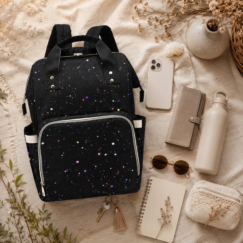 May include: A black backpack with a galaxy design, speckled with colourful dots. The image also shows a phone, wallet, sunglasses, notebook, water bottle, and a small purse, all arranged on a neutral surface.