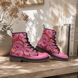 Op de afbeelding: Een paar enkelhoge veterschoenen met een herhalend patroon van roze geglazuurde donuts met kleurrijke hagelslag. De laarzen hebben zwarte veters, zwarte oogjes, een effen roze neus en een dikke, donkerbruine rubberen zool met een grof profiel.