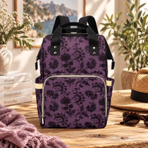 Puede incluir: Una mochila morada con un patr&oacute;n floral de damasco negro, asas superiores negras y correas de hombro negras. La bolsa incluye un bolsillo frontal con cremallera y herrajes dorados, bolsillos laterales y una forma rectangular estructurada.