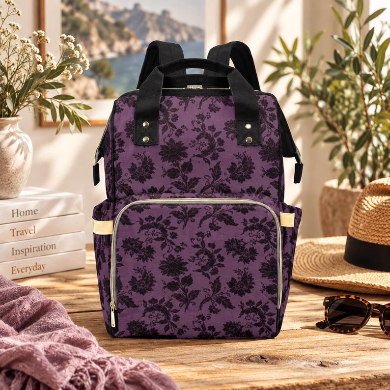 Puede incluir: Una mochila morada con un patr&oacute;n floral de damasco negro, asas superiores negras y correas de hombro negras. La bolsa incluye un bolsillo frontal con cremallera y herrajes dorados, bolsillos laterales y una forma rectangular estructurada.