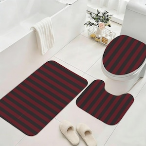 May include: A three-piece bathroom mat set featuring a dark red and black vertical striped pattern. The set includes a rectangular floor mat, a U-shaped contour mat for the toilet base, and a matching cover for the toilet lid.