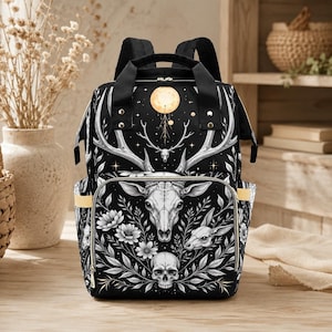 Puede incluir: Mochila negra con diseño de calavera de ciervo blanco, floral y lunar. La mochila tiene un asa superior, correas ajustables y bolsillos laterales. El diseño incluye una cabeza de ciervo, una calavera y una luna llena.