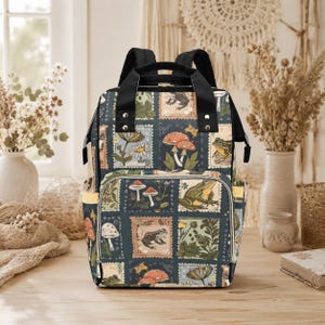 Puede incluir: Una mochila de color verde azulado oscuro con un patrón repetido de ilustraciones estilo sello postal con setas, ranas y flores. La mochila tiene correas negras y un asa superior, con detalles amarillos en los bolsillos laterales.