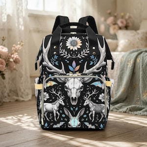Può includere: Zaino nero con un design floreale e teschio di cervo. Lo zaino presenta una grande immagine centrale di un teschio di cervo, circondato da motivi floreali e di corna. Le tasche laterali hanno finiture gialle. La borsa ha cinghie nere e una maniglia superiore.