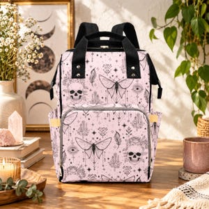 May include: A light pink backpack featuring a black line art pattern of skulls, moths, crystals, and stars. The bag has black top handles, adjustable shoulder straps, a front zippered pocket, and side pockets with gold-toned metal hardware.
