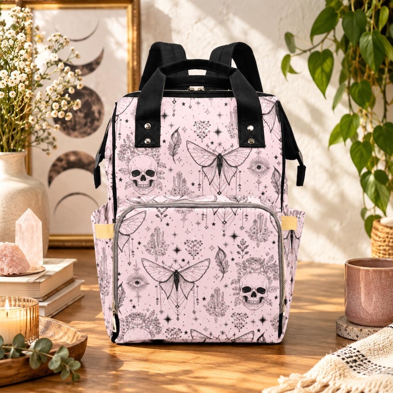 May include: A light pink backpack featuring a black line art pattern of skulls, moths, crystals, and stars. The bag has black top handles, adjustable shoulder straps, a front zippered pocket, and side pockets with gold-toned metal hardware.