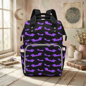 May include: A black backpack featuring a repeating pattern of purple bats. The bag includes a large main compartment, a front zippered pocket, two top carry handles, and adjustable shoulder straps with silver-toned metal hardware accents.