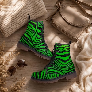 May include: A pair of black and neon green zebra print combat boots with black laces. The boots are displayed on a wooden surface with a beige sweater, sunglasses, and a purse.