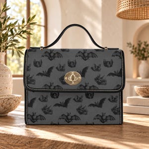 May include: A rectangular handbag featuring a dark gray surface with a repeating pattern of black bats in various flight positions. The bag has a black top handle, black trim, and a gold-toned metal turn-lock closure centered on the front flap.