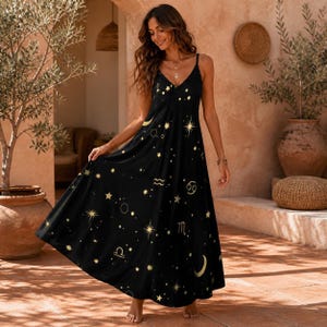 Könnte beinhalten: Ein schwarzes Maxikleid mit V-Ausschnitt und dünnen Spaghettiträgern. Der Stoff ist mit einem goldenen Himmelsdruck verziert, der aus verschiedenen Sternen, Mondsicheln und Tierkreiszeichen besteht, die über die gesamte Stoffoberfläche verteilt sind.