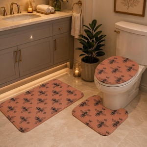 May include: A bathroom set featuring a coral-colored rug, toilet seat cover, and contour rug, all adorned with a repeating black bee pattern. The set includes a rectangular rug, a toilet seat cover, and a U-shaped rug.