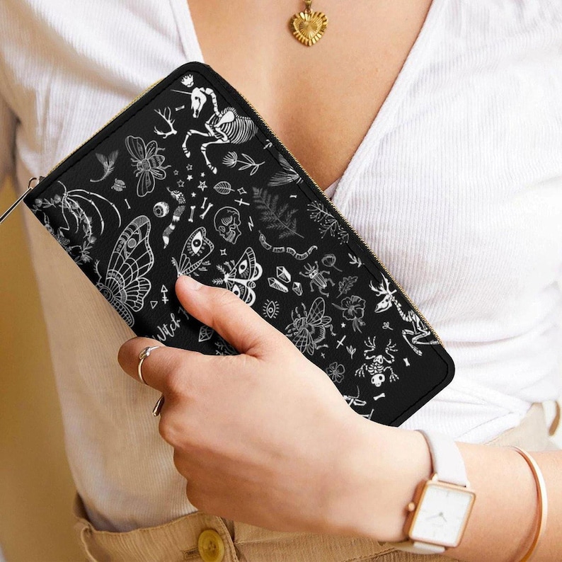 A person wearing a white top holds a black zippered wallet with a white skeleton and botanical design.