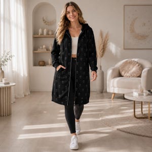 May include: A black hooded duster coat with a skull pattern. The coat has a full-length zip and pockets. The person is wearing black leggings and white trainers.