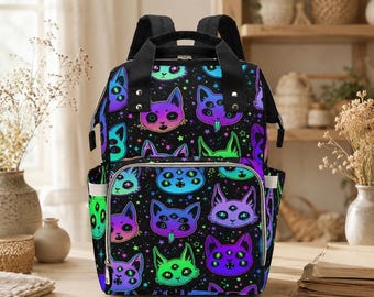 May include: A black backpack featuring a repeating pattern of colorful, multi-eyed cat faces in shades of neon green, purple, and blue, accented with small stars. The bag includes black top handles, adjustable shoulder straps, and a front zipper pocket.