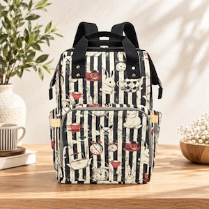 May include: A backpack featuring a black and white vertical striped pattern with a repeating print of white rabbits, red and white teacups, teapots, and clocks. The bag has black top handles, adjustable shoulder straps, and a front zippered pocket.