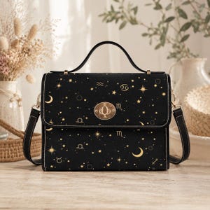Könnte beinhalten: Schwarze Handtasche mit einem Himmelsdesign mit goldenen Sternen, Monden und Tierkreiszeichen. Die Tasche hat einen oberen Griff, einen Schultergurt und einen goldenen Verschluss. Die Nähte sind weiß.