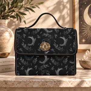 May include: A black rectangular handbag featuring a repeating pattern of white crescent moons and leafy branches. The bag has a single top handle, a gold-toned metal twist-lock closure on the front flap, and black piping along the edges.