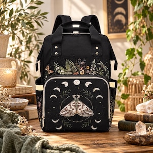 May include: A black backpack featuring a printed design of a moth centered within a lunar cycle. The front pocket displays a botanical arrangement of ferns and wildflowers. The bag includes top handles, adjustable shoulder straps, and side pockets.