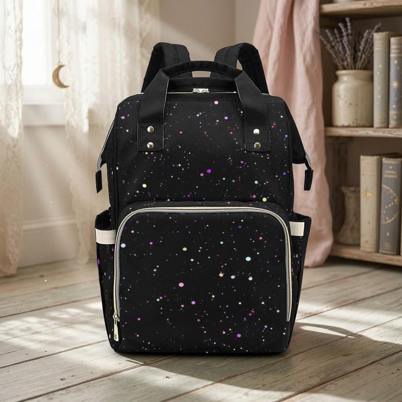May include: Black backpack with a galaxy-themed pattern of colourful dots. The bag has a large front pocket with a white trim and side pockets. It features a top handle and adjustable shoulder straps.
