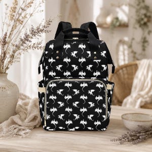 Puede incluir: Mochila negra con un estampado de dragones blancos. La bolsa tiene múltiples compartimentos, un asa superior y correas ajustables. El bolsillo delantero tiene una cremallera dorada y ribete beige. La bolsa está sobre una superficie de madera.