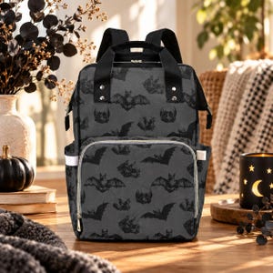 May include: A dark grey backpack featuring an all-over print of black bats in various flight positions. The bag includes black top handles, adjustable shoulder straps, a front zippered pocket, and side pockets with white trim and silver zips.