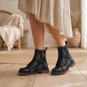 May include: Black lace-up ankle boots featuring a repeating pattern of gold-colored moths and small stars. The boots have a rounded toe, a sturdy sole with a low heel, and black laces threaded through metal eyelets along the front shaft.