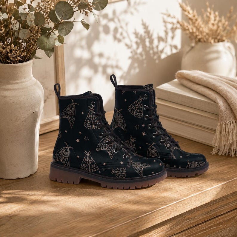 May include: A pair of black ankle boots featuring a repeating pattern of light-colored moths and small stars. The boots have black laces, metal eyelets, and a thick, textured sole. The design covers the entire upper portion of the footwear.