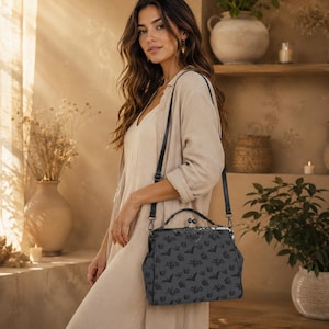 May include: A black handbag with a bat pattern, featuring a clasp closure and a shoulder strap. The bag is held by a person wearing a beige outfit. The background includes plants, candles, and a shelf.