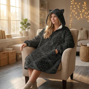 May include: A dark gray hooded blanket with a black floral pattern. The blanket has a zipper closure, front pockets, and cat ears on the hood. The person is sitting in a beige armchair.