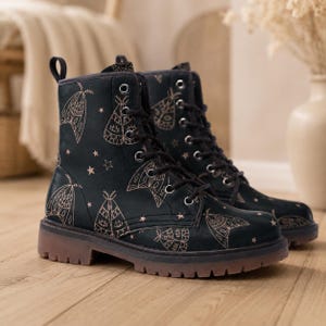 May include: A pair of black lace-up ankle boots featuring a repeating pattern of light-colored moth illustrations and small stars. The boots have black laces, silver-toned metal eyelets, a pull tab at the heel, and a thick, translucent brown lug sole.