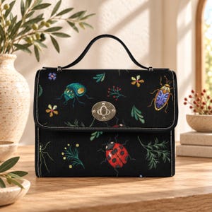 Könnte beinhalten: Eine schwarze, strukturierte Handtasche mit einem Tragegriff und einem silberfarbenen Drehverschluss aus Metall. Der Außenstoff ist mit einem bunten Stickmuster aus verschiedenen Insekten, darunter ein Marienkäfer, und kleinen Blumenmotiven verziert.